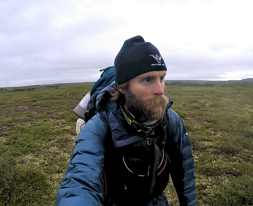 Adam Shoalts hikes with a large backpack on. The landscape is flat but filled with moss and rocks.