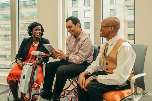 Three people sit next to each other. One person sitting in the middle uses and assistive chair with wheels.