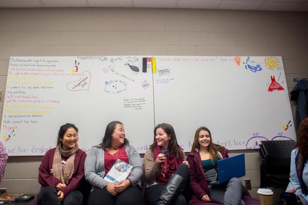 Four women laugh while sitting on a couch in front of a white board.