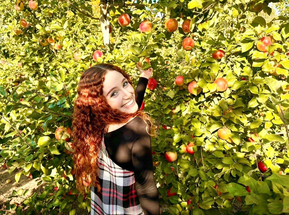 Juliette smiles while reaching out to pick an apple in an apple orchard.